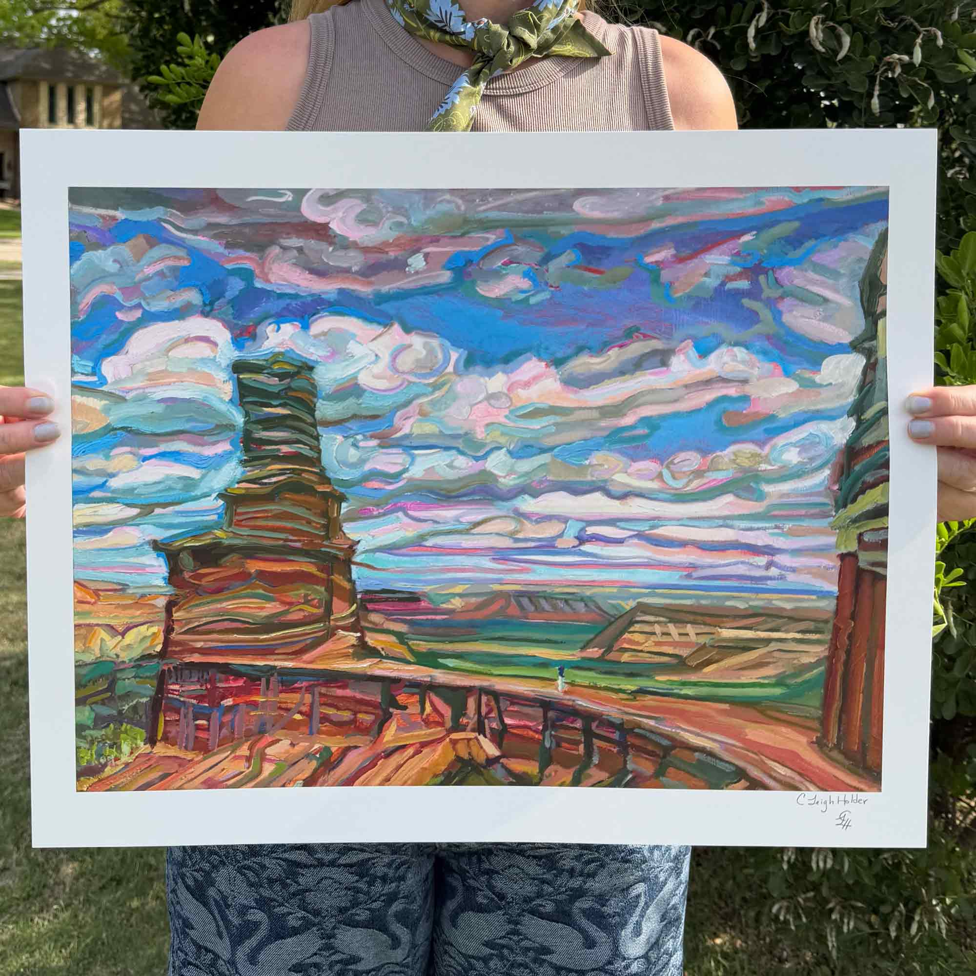 Arist holding a colorful landscape painting of Palo Duro Canyon with a natural bridge and cotton candy Texas clouds with blue sky.