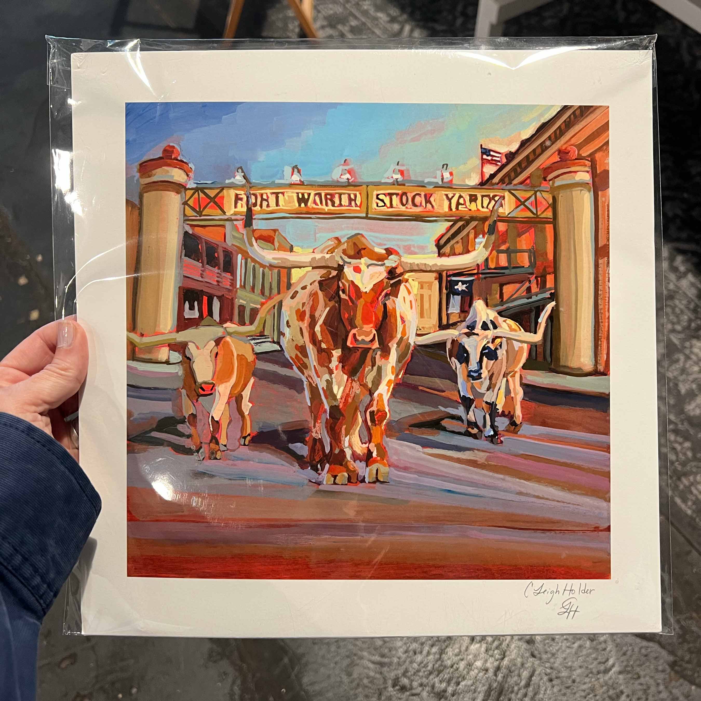 Painting of Fort Worth Stock Yards with longhorn cattle, held in a clear plastic sleeve. Art print size is 12x12.