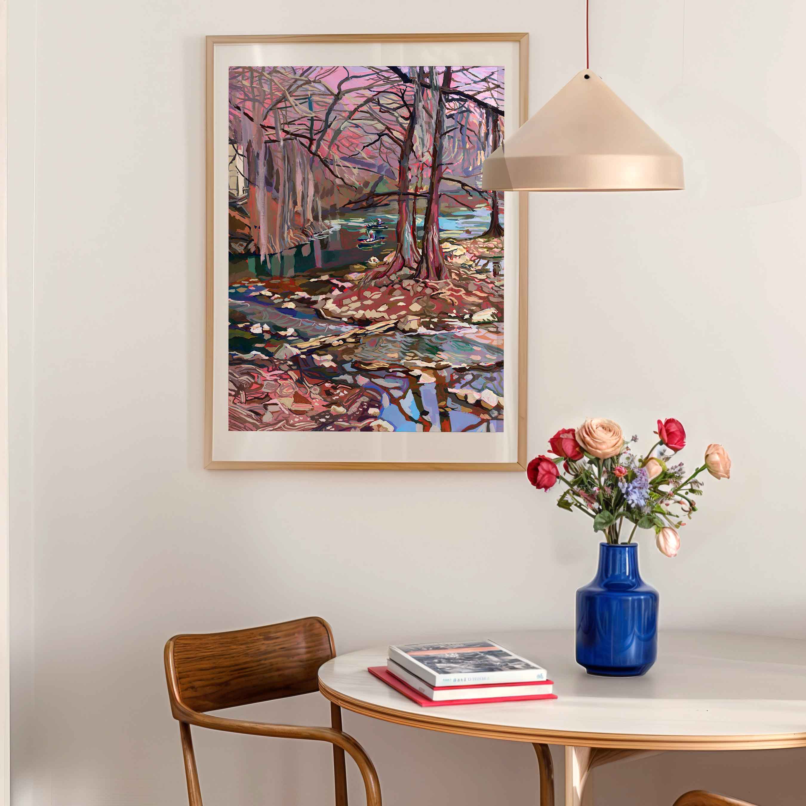 Framed abstract painting of river on a wall in a retro kitchen above a table with a vase of flowers and books.