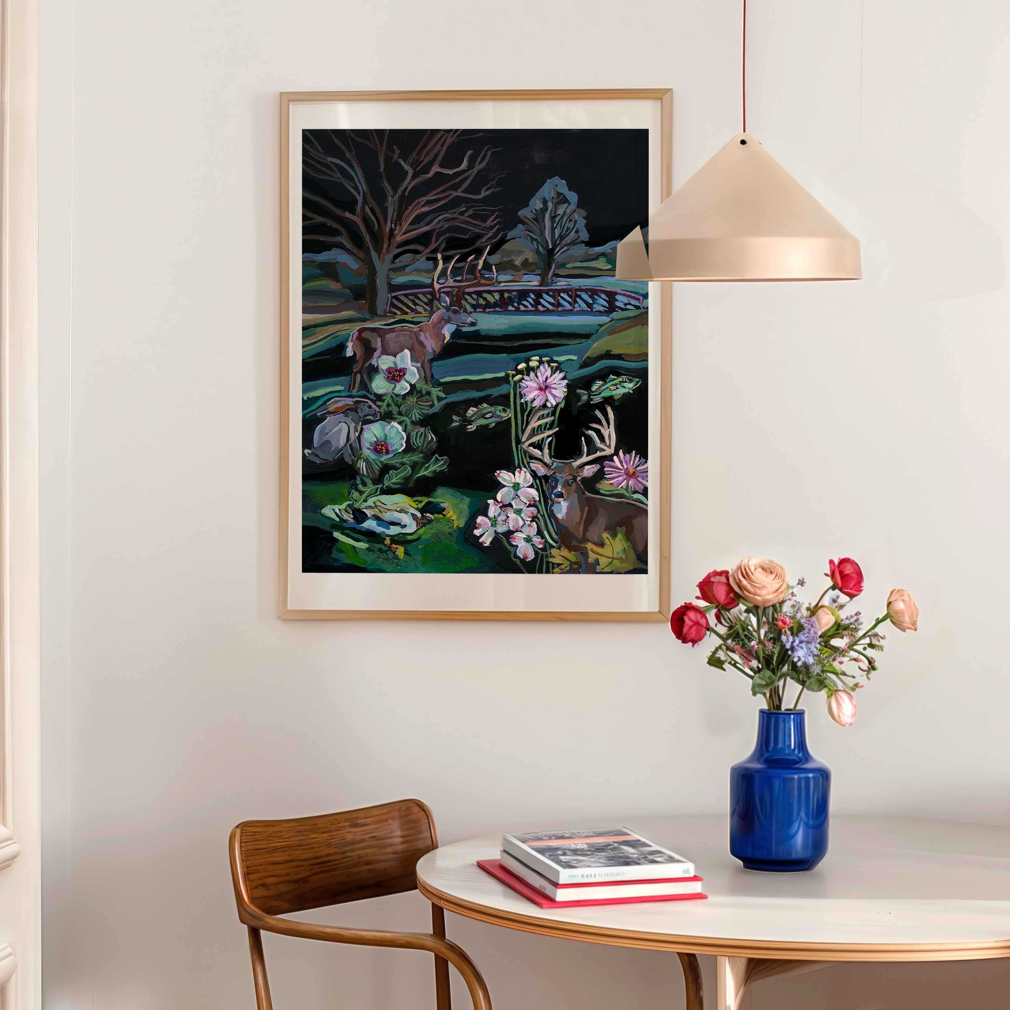Painting of whitetail deer, duck, bass fish and nature scene on Texas river in New Braunfels, Texas on a blue fabric background.Framed artwork on a vintage kitchen wall above a table with a vase of flowers and books.